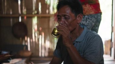 A humble old Asian Lahu sits on the floor drinking tea from a crystal glass in his simple rural house made of reeds and wood - 4K Horizontal