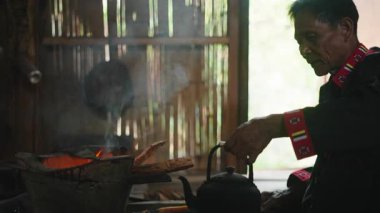 A man in traditional clothes carefully tends a small fire to prepare tea in a rustic setting - 4K Horizontal