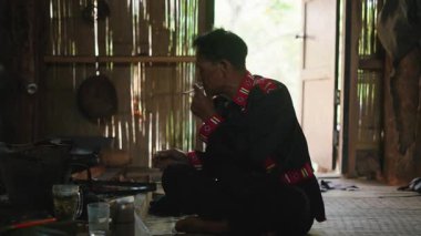 An Asian man from the Lahu tribe smokes a cigarette while preparing a meal in a traditional hut with bamboo walls - 4K Horizontal