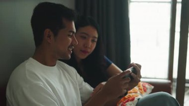 Happy young asian couple sitting on the sofa playing video games on a mobile phone together