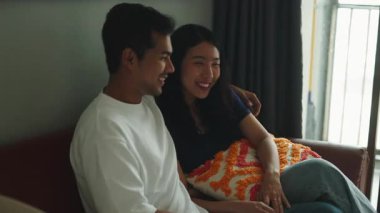 Happy young asian couple sitting on the sofa, talking and laughing in a cozy living room