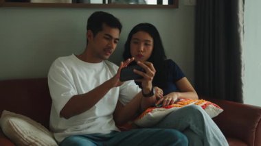 Happy young asian couple sitting on a sofa watching social media content on a mobile phone