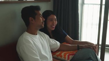 Affectionate young asian couple sitting on sofa, holding hands, talking, and laughing together