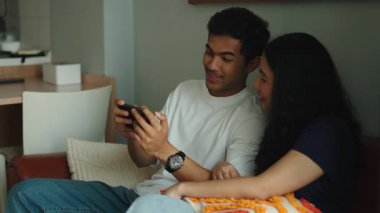 Cheerful asian man and woman sitting on a couch watching videos and playing games on a mobile phone