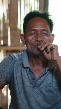 Yaşlı bir Asyalı, tahta bir kulübede bağdaş kurup oturmuş sakince el ile sarılmış bir sigara içiyor.