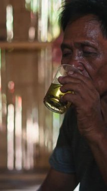 Yaşlı, mütevazı bir Asyalı Lahu, sazlıktan ve ahşaptan yapılmış sade kırsal evinde kristal bir bardaktan çay içerek yerde oturur.