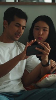 Mutlu genç Asyalı çift kanepede oturuyor, gülümsüyor ve evde birlikte cep telefonu kullanıyorlar.