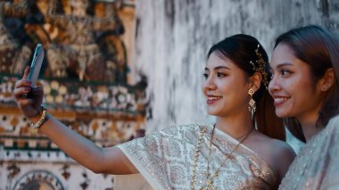 Geleneksel Thai elbiseli iki gülümseyen kadın kültürlerini ve dostluklarını kucaklıyor, akıllı telefonlarıyla anları yakalıyorlar.