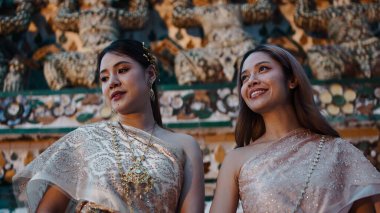 Geleneksel Tayland kostümü giymiş iki arkadaş antik bir tapınağın önünde gülüyor, bir festivali kutluyorlar.