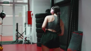 Kulaklıklı genç bir kadın modern bir dövüş salonunda tekmeleyip yumrukluyor.