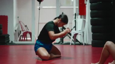 Kendini işine adamış kadın dövüş sanatları ustası kırmızı bir paspasın üzerine diz çöküp kendini korumak için ellerini dikkatlice sarıyor.