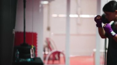 Kickboks antrenmanı yapan kararlı genç bir kadın spor salonunda ağır bir torbaya yumruk atıyor. 4K yatay.