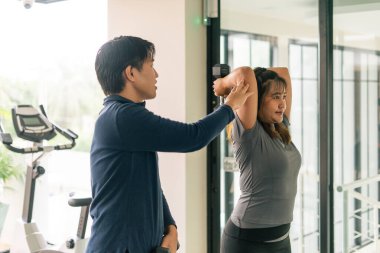 Kişisel antrenör, genç bayana dambıl egzersizinde yardımcı oluyor, egzersiz sırasında doğru formu sağlıyor.