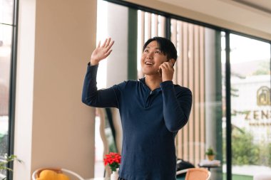 Gülümseyen Asyalı genç adam telefonda konuşuyor, el sallıyor, modern iç mekanda jest yapıyor.