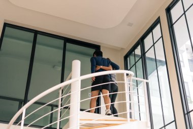 Genç çift kucaklaşıyorlar, modern, aydınlık bir evde temiz, sarmal bir merdivende yürüyorlar.