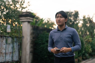 Genç Asyalı bir adam dışarıda yürürken akıllı bir telefon tutuyor, ileri bakıyor, düşünüyor, seyahat ediyor.