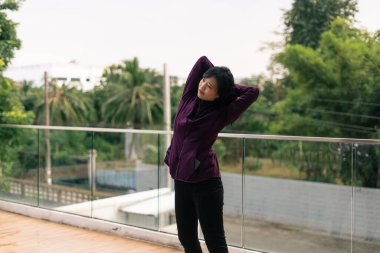 Asyalı kadın ısınıyor, yan vücudunu modern bir balkon korkuluğuna geriyor.