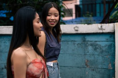 İki Asyalı kadın, yıpranmış mavi bir duvarın yanında yürürken neşeli bir sohbetin tadını çıkarıyorlar.
