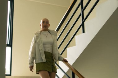 Modern bir merdivenden inen kel Asyalı kadın, gücünü ve benzersiz kimliğini ifade ediyor.