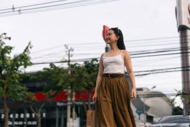 Bir şehir caddesinde elinde alışveriş çantası, yürürken gülümseyen genç bir kadın.