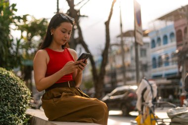 Gün batımında bir şehir sokağında, duvarda oturan bir kadın telefonuyla sosyal medyayı kaydırıyor.