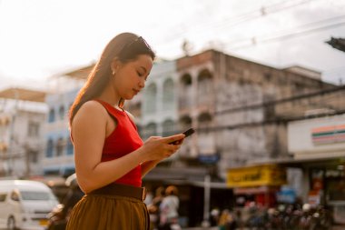 Genç Asyalı bir kadın şehir caddesinde cep telefonuyla iletişim kuruyor ve günlük hayatın tadını çıkarıyor.