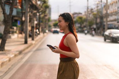 Gülümseyen genç bir kadın şehir sokağında duruyor, başka tarafa bakıyor, cep telefonu kullanıyor.