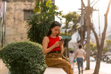 Genç bir kadın dışarıda oturuyor, cep telefonu kullanıyor, günlük yaşam tarzını temsil ediyor.