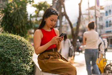 Duvarda oturan genç bir kadın, modern bir şehir caddesinde telefonuna sarılmış.
