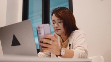 Masasında dizüstü bilgisayarla otururken akıllı telefonunu kullanan gözlüklü, olgun Asyalı bir iş kadını.