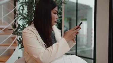 Modern bir iç mekanda rahat bir şekilde otururken genç profesyonel bir kadın cep telefonunu kaydırıyor.