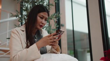 Pencerenin yanındaki merdivenlerde oturan zarif bir kadın beklerken cep telefonuyla mesaj atıyor.