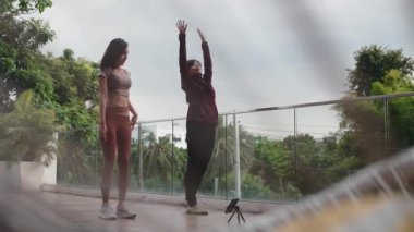 Çevrimiçi bir dersi takip eden iki genç Asyalı kadın modern bir binanın balkonunda egzersiz yapıyor ve esniyorlar.