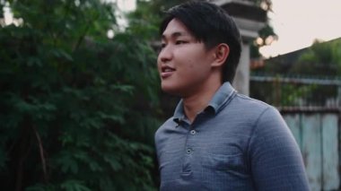 Yakışıklı Asyalı bir adam birini bekliyor ve bahçede dikilirken gülümsüyor.