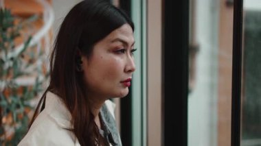 Kablosuz kulaklık takan ve yağmurlu bir sonbahar gününde pencereden dışarı bakan düşünceli Asyalı kadın.