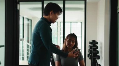 Spor salonunda akıllı telefon uygulaması kullanan bir bayan müşteriyle kişisel antrenör gelişimi tartışıyor.