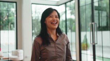 Neşeli Asyalı kadın gülümsüyor, el sallıyor ve oturma odasında arkadaşlarına doğru yürüyor.