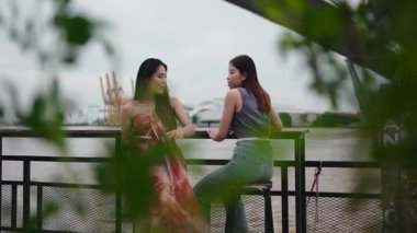 Genç Asyalı bayan arkadaşlar nehir kenarındaki bir kafede gülüyor ve akıllı telefon kullanıyorlar.