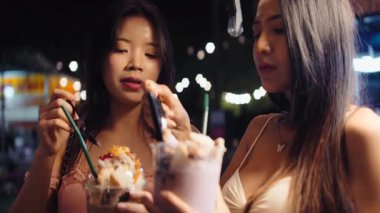 İki Asyalı genç arkadaş şehirdeki açık hava gece pazarında lezzetli tatlılar yiyorlar.