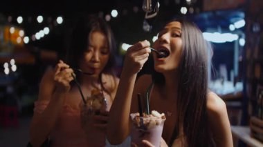 İki mutlu Asyalı kadın gece canlı bir cadde yiyecek pazarında lezzetli bir tatlı yiyorlar.