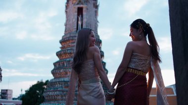İki Taylandlı kadın el ele tutuşuyor, birbirlerine bakıyor, Wat Arun 'a gülümsüyorlar. Loi Krathong gününü kutluyoruz.