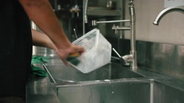 Erkek mutfak işçisi, profesyonel paslanmaz çelik lavaboda büyük bir plastik gıda kabını yıkıyor.