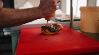 Şefin elleri kırmızı kesim tahtasındaki sığır burgerine şişeden sos sıkıyor.