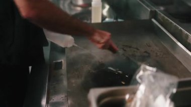 Çalışkan aşçı, dumanı üstünde bir ızgarayı suyla temizliyor ve ticari bir mutfakta kazıyıcı kullanıyor.