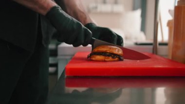 Siyah eldiven giyen erkek aşçı kırmızı kesme tahtasındaki yeni yapılmış hamburgeri kesiyor.