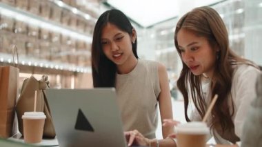İki Asyalı genç bayan arkadaş bir kafede dizüstü bilgisayar kullanırken konuşup gülüyorlar.