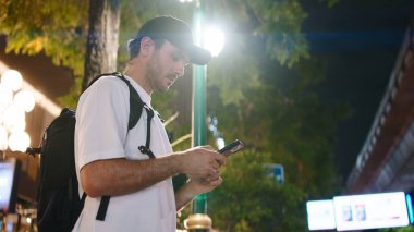 Sırt çantalı bir adam gece şehir yolculuğu sırasında aydınlık bir caddede telefonunu kontrol ediyor.