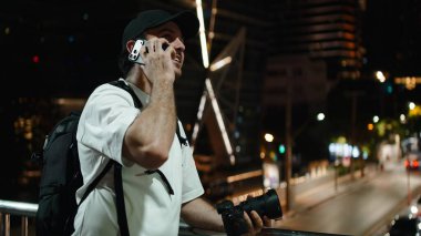 Erkek fotoğrafçı akıllı telefondan konuşuyor, elinde kamera, şehir manzarasını inceliyor.