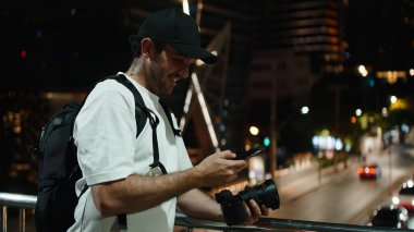 Sırt çantalı ve kameralı gülümseyen adam gece akıllı telefon ekranını tarıyor.