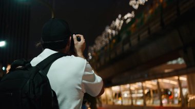 Kameralı ve sırt çantalı fotoğrafçı, gece canlı şehir ışıklarını belgelemiş, varış noktasını araştırıyor.